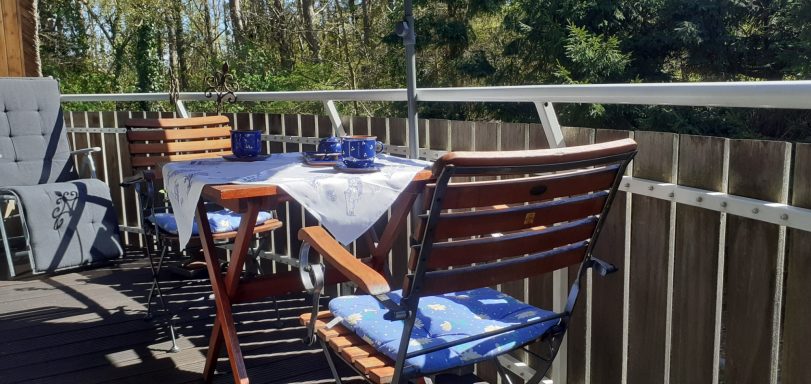 Holztisch mit blauem Tischtuch und Stühlen auf einem sonnigen Balkon umgeben von Bäumen.