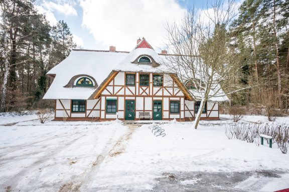 Holzhaus im Schnee, umgeben von Bäumen, mit einem roten Dach und grünen Türen.